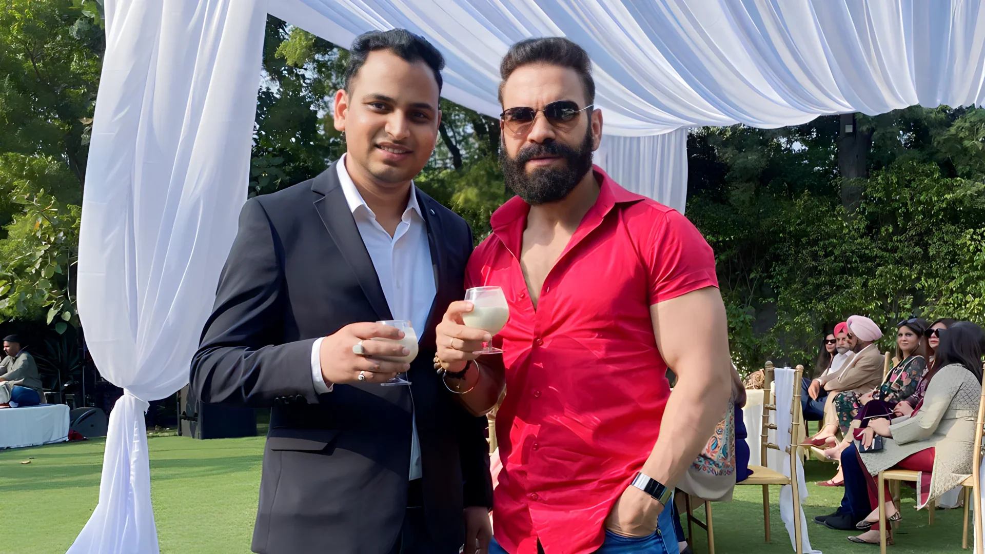 Lithvik Mukesh Sharma is photographed. Two men stand outdoors on a lawn under a white fabric canopy. Lithvik Mukesh Sharma, the younger man, wears a black suit. The older man wears a bright red/pink shirt